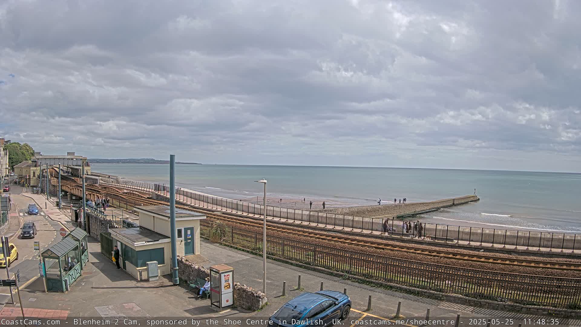 A coastal railway line runs alongside a beach with a few people walking on the sand under a partly cloudy sky.