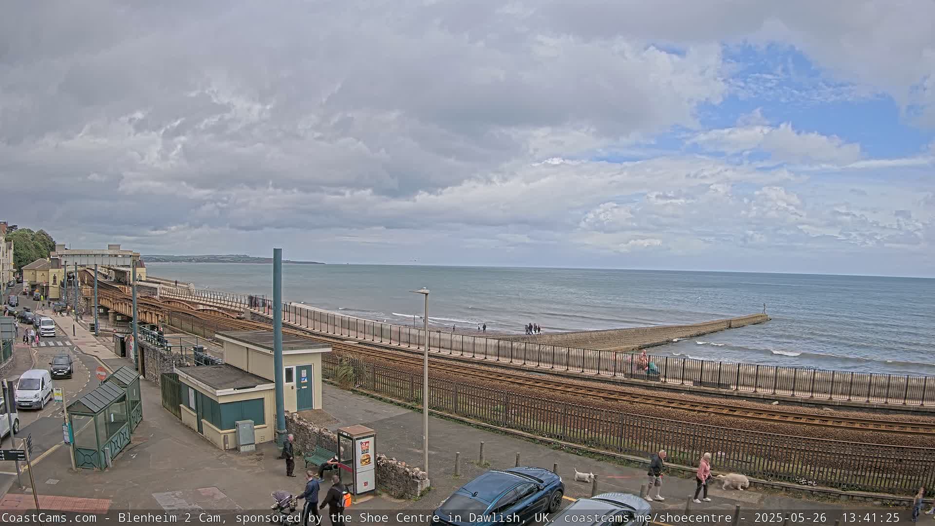 A mostly cloudy day shows a seaside town with train tracks running alongside a beach and a breakwater extending into a wavy ocean.