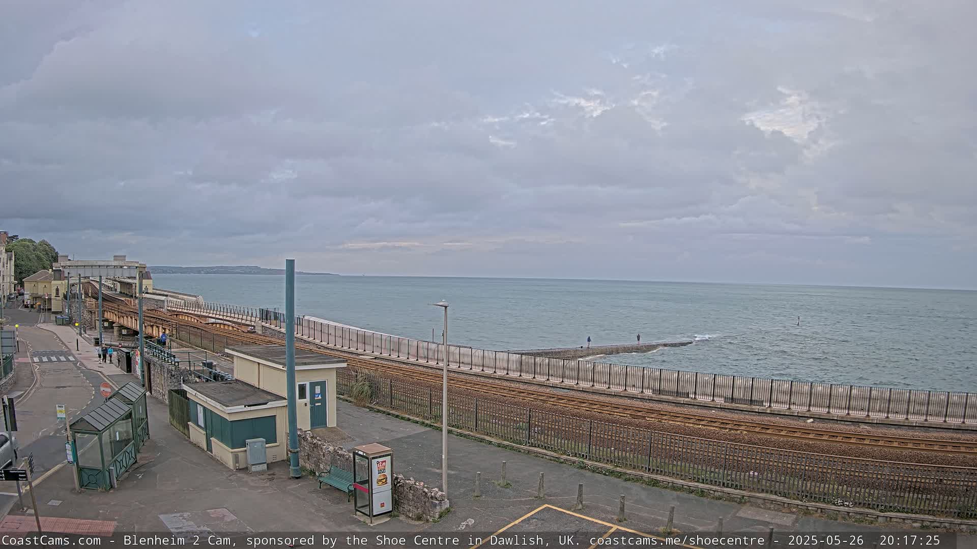 A train track runs alongside a sea wall bordering a calm, overcast ocean.