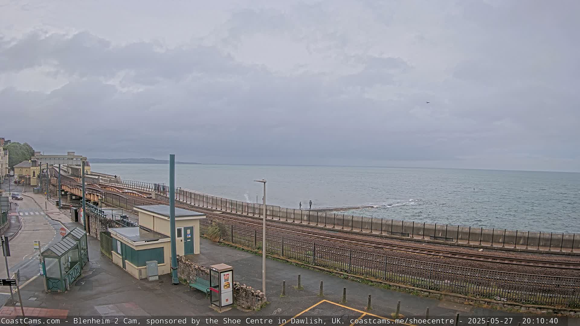 A coastal train track runs alongside a sea wall, with a calm sea and overcast sky in the background.
