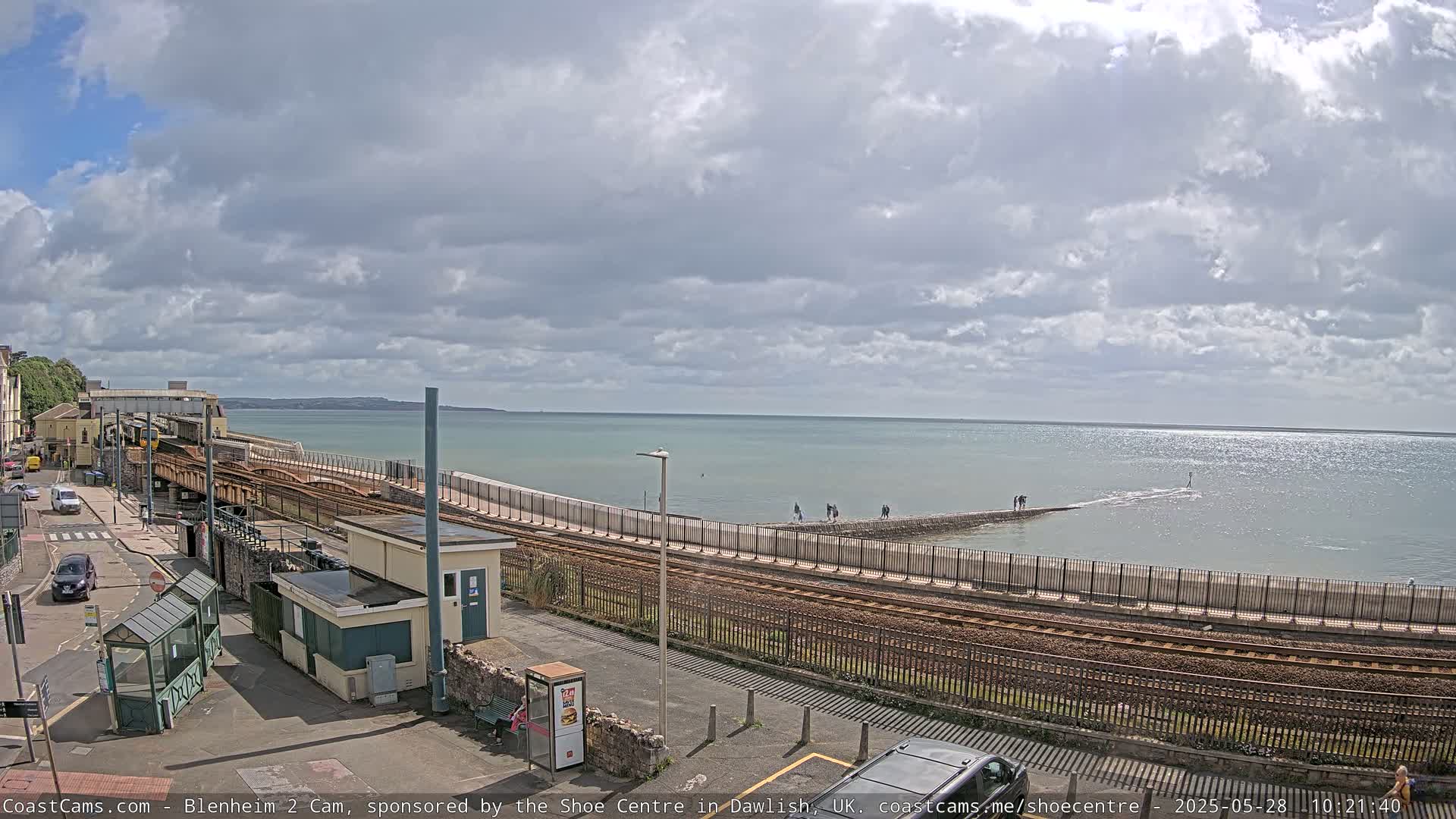 A mostly cloudy day overlooks a coastal train line, a sea wall with people walking on it, and a calm body of water extending to the horizon.