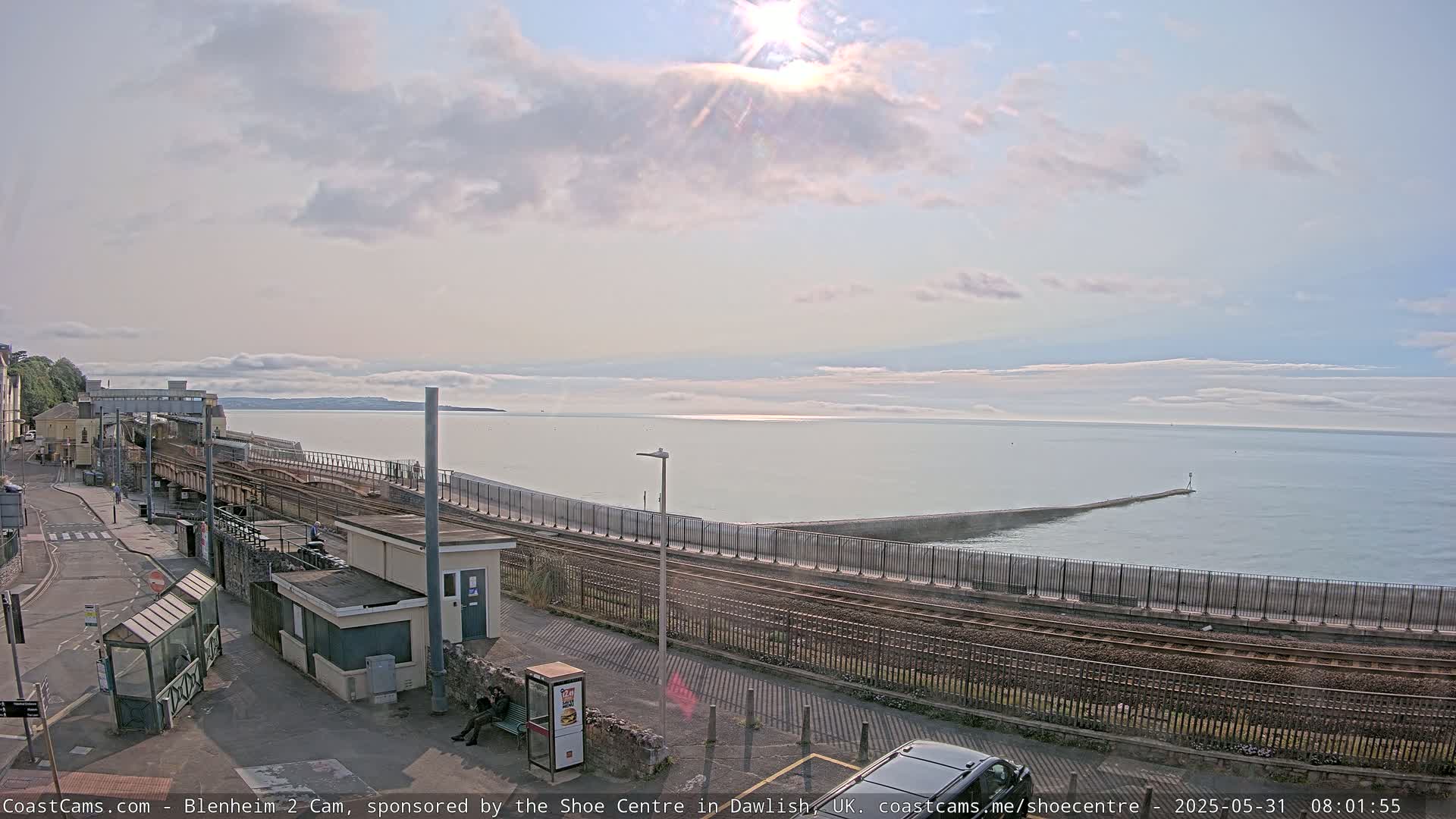 A sunny day shows a train station next to a calm sea with a breakwater, and railroad tracks running along the water's edge.