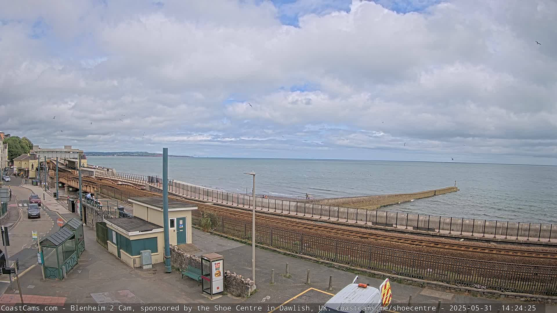 A train track runs alongside a beach and sea wall, with a cloudy sky overhead and a calm sea.