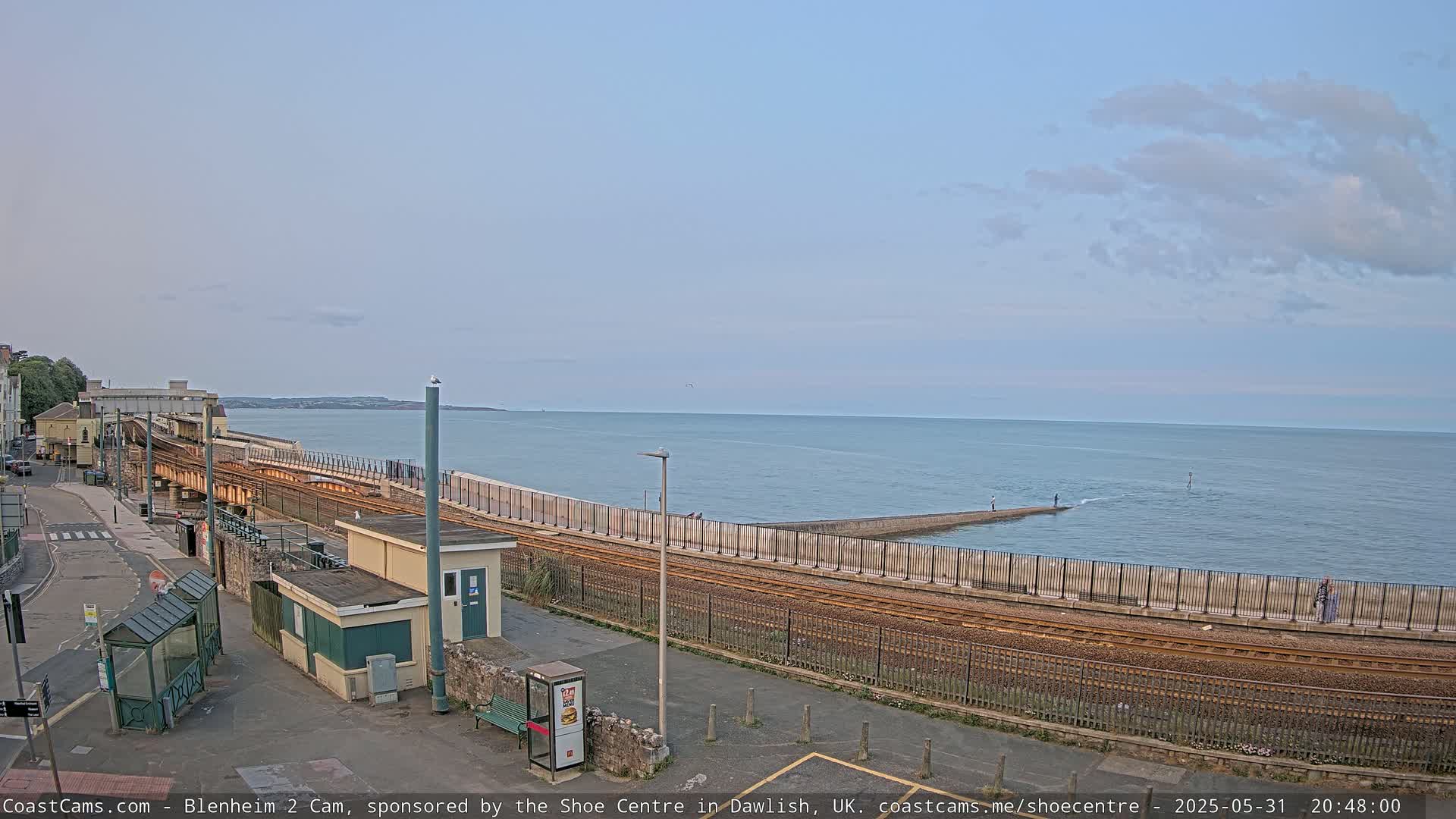 A coastal train track runs alongside a seawall separating a road from a calm ocean under a partly cloudy sky.