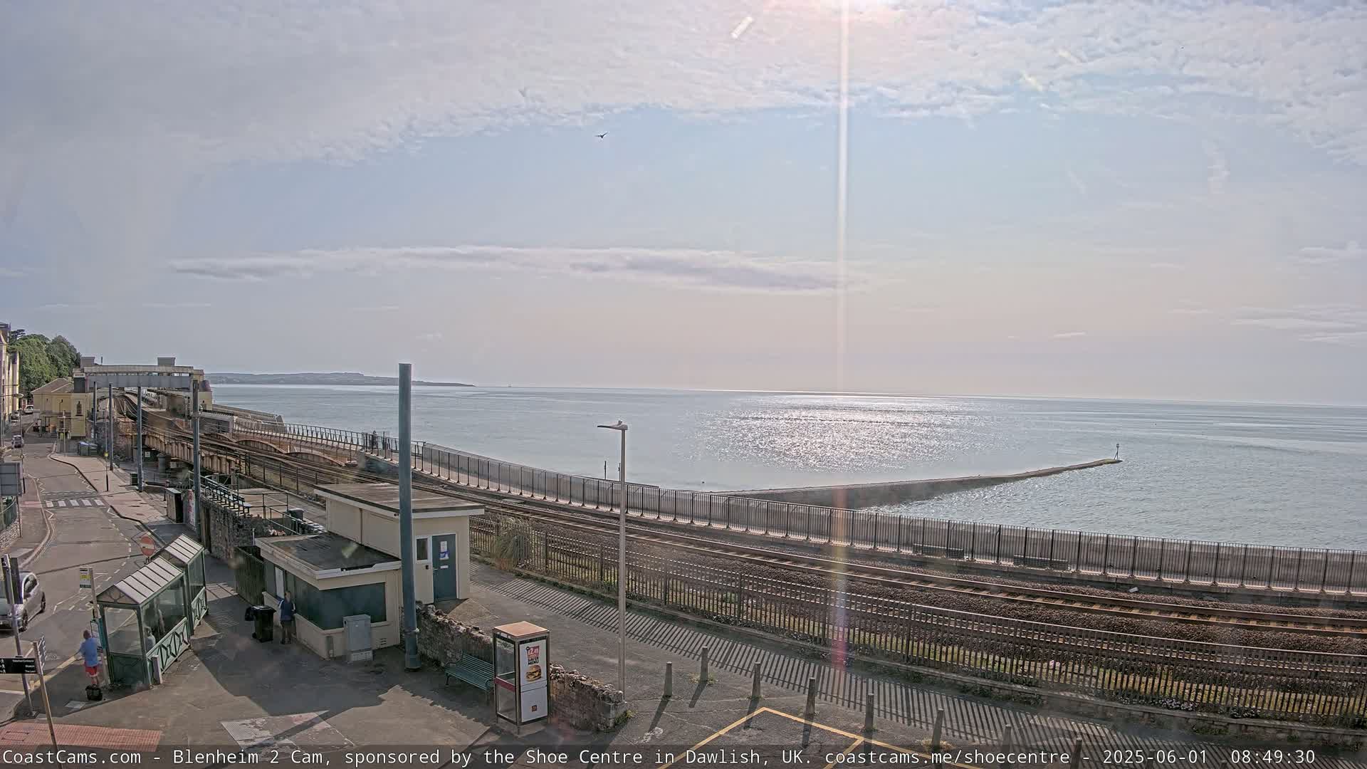 A train track runs alongside a calm sea, with a breakwater extending into the water under a partly sunny sky.