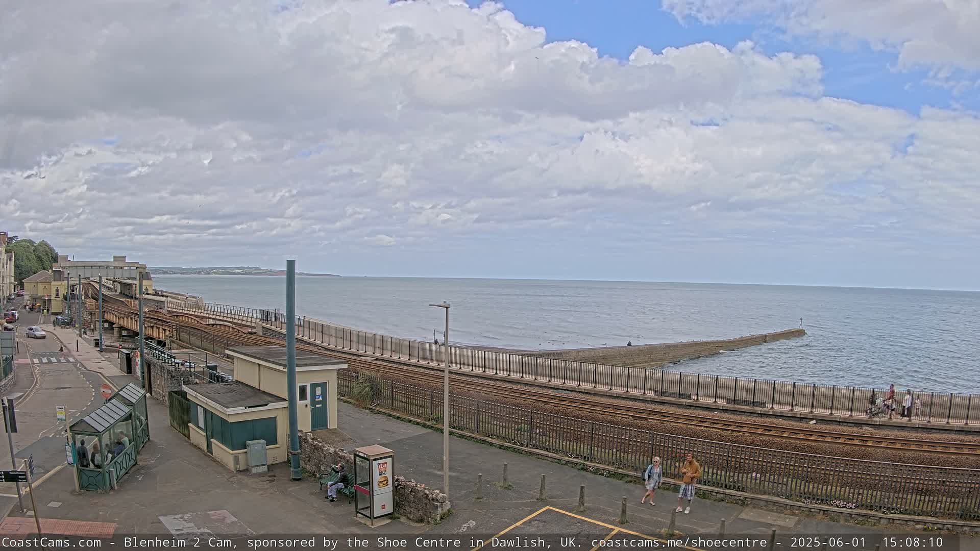A train track runs alongside a seawall, next to a calm ocean under a partly cloudy sky.