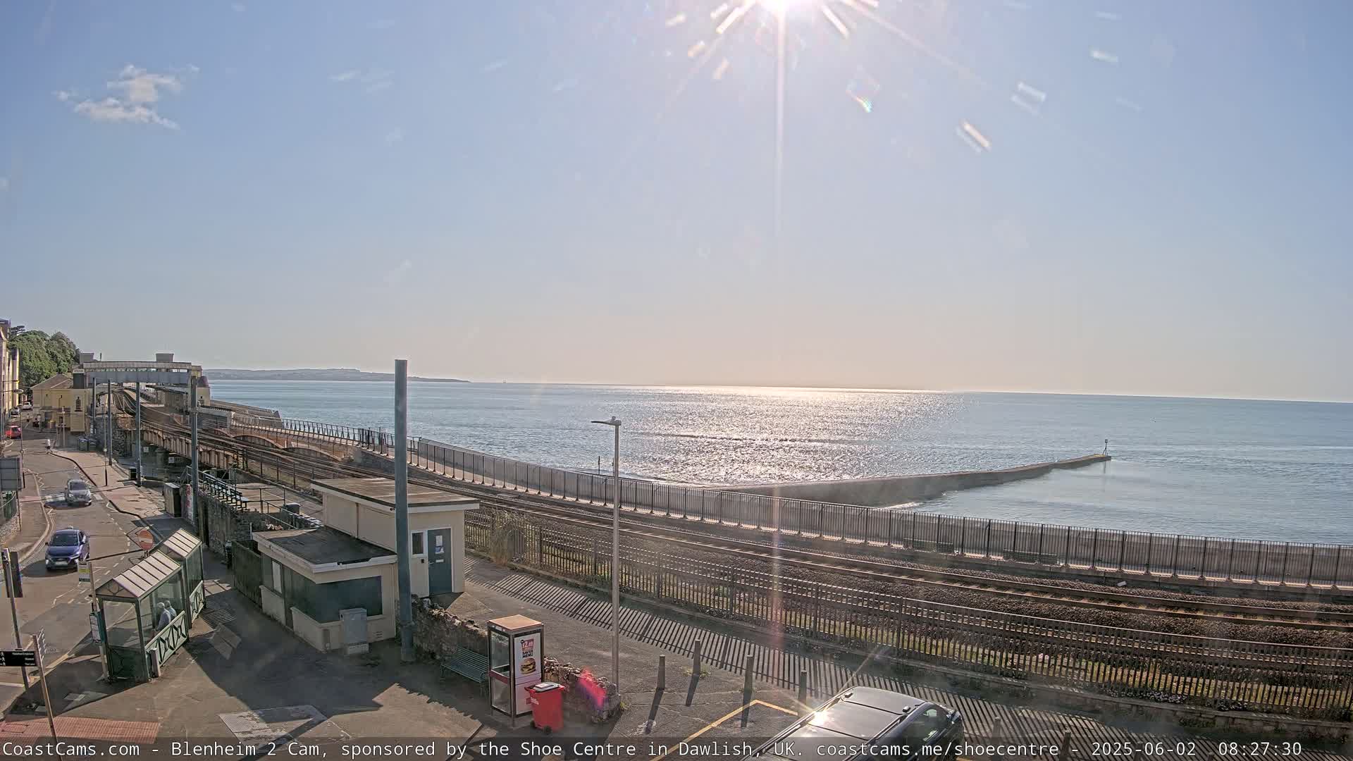 A sunny day at a seaside train station, with calm ocean waters and a breakwater extending into the sea.