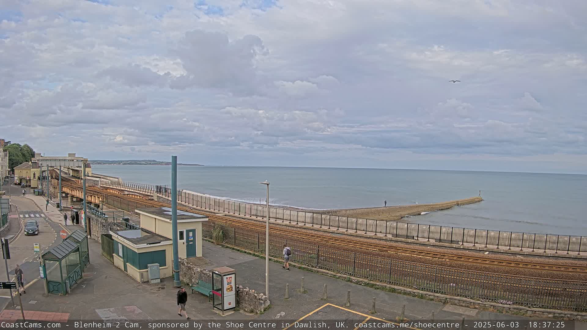 A coastal railway line runs alongside a beach and sea wall under a partly cloudy sky.