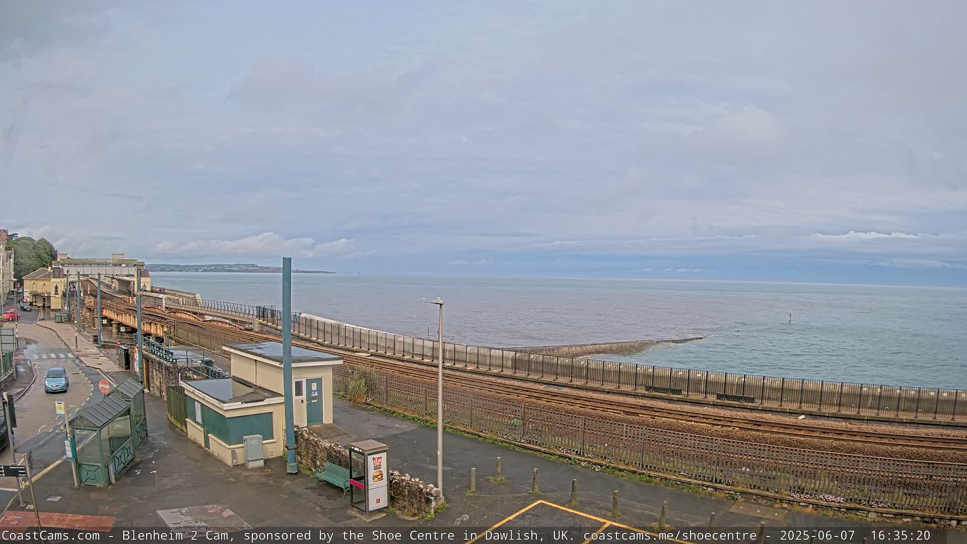 A train track runs along a seawall next to a calm, overcast ocean.