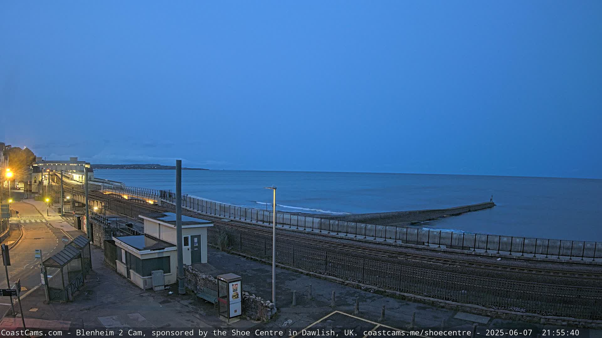 A coastal train station at twilight, with a calm sea and a breakwater extending into the water.
