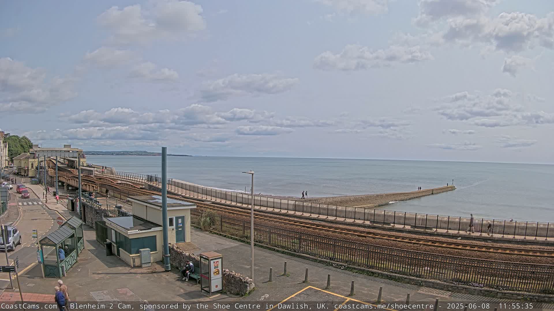 A seaside train station with railway tracks leading to a breakwater, under a partly cloudy sky.