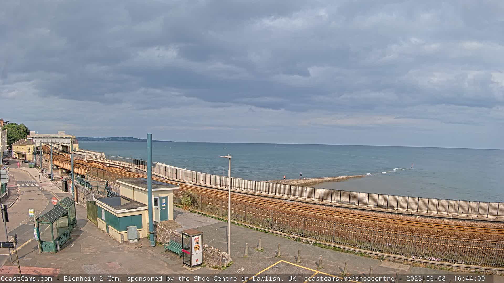 A coastal train track runs alongside a sea wall, with a calm sea under a cloudy sky.