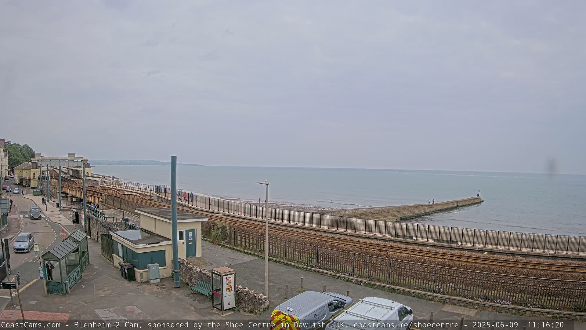 A coastal railway line runs alongside a beach and calm sea under an overcast sky.