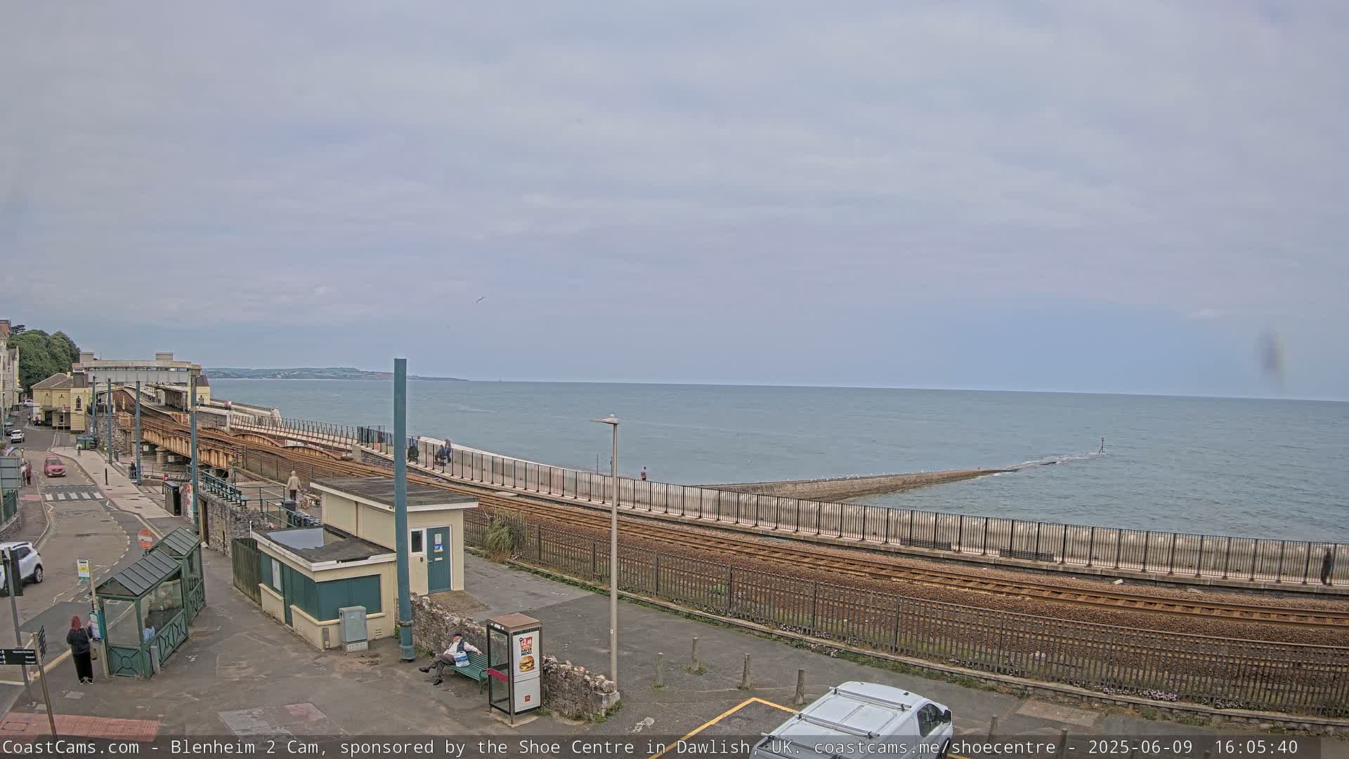 A coastal railway line runs alongside a sea wall, with a calm sea and overcast sky in the background.