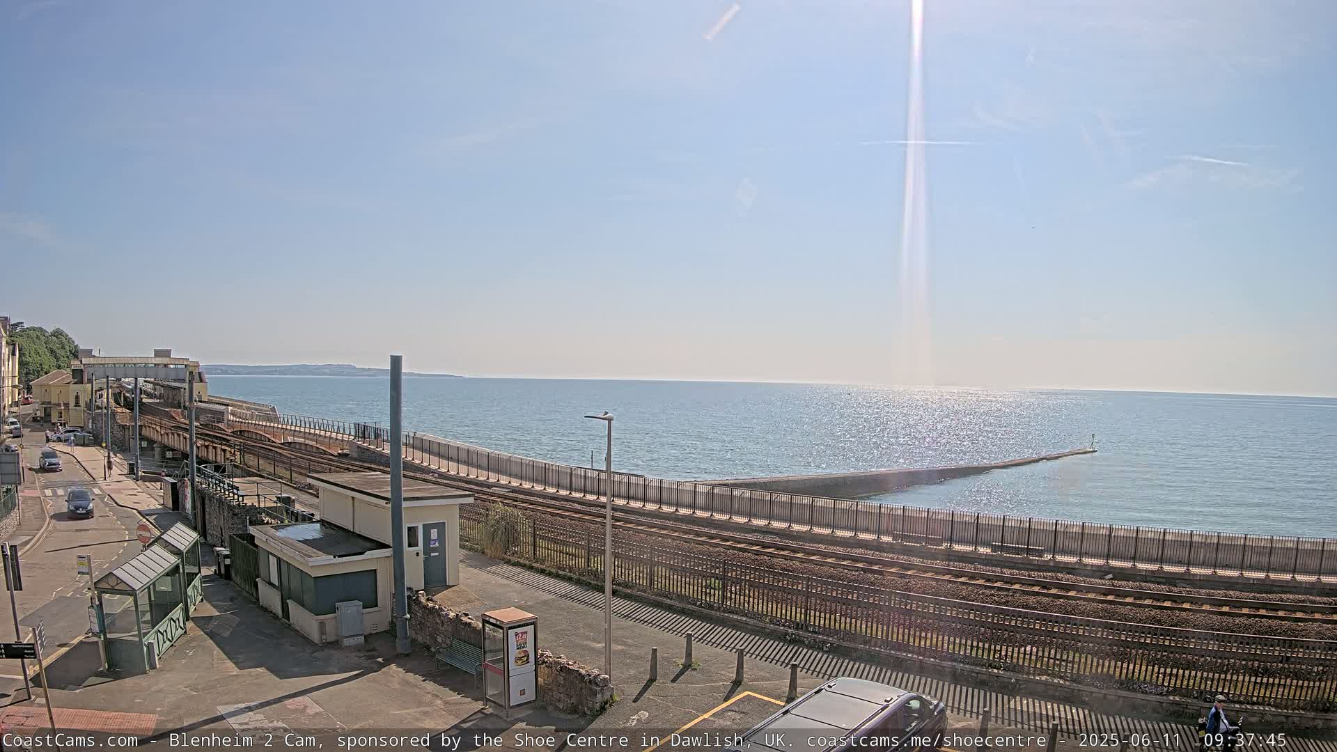A sunny seaside scene shows a train track running alongside a sea wall and out to a breakwater, with a calm ocean beyond.