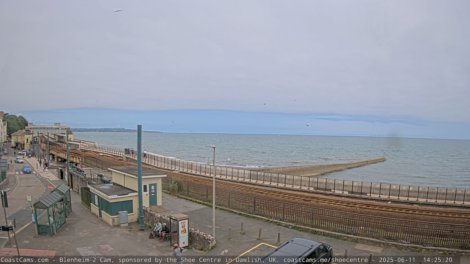 A seaside train track curves alongside a seawall separating a calm ocean from a paved area with small buildings and people.