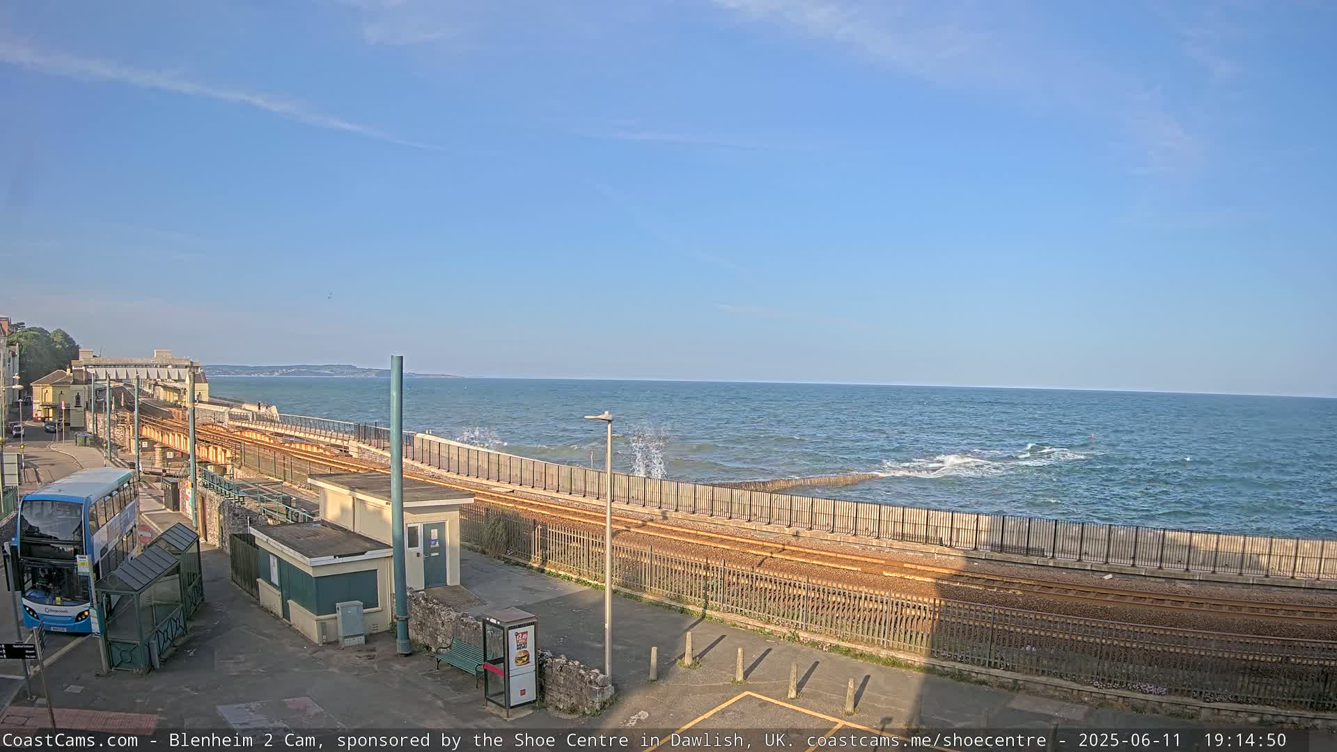 A seaside train track runs alongside a wall protecting the shore from moderately rough ocean waves under a mostly clear sky.