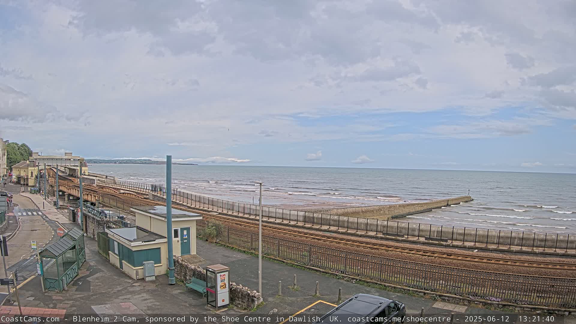A seaside train track runs alongside a beach with gentle waves under a partly cloudy sky.