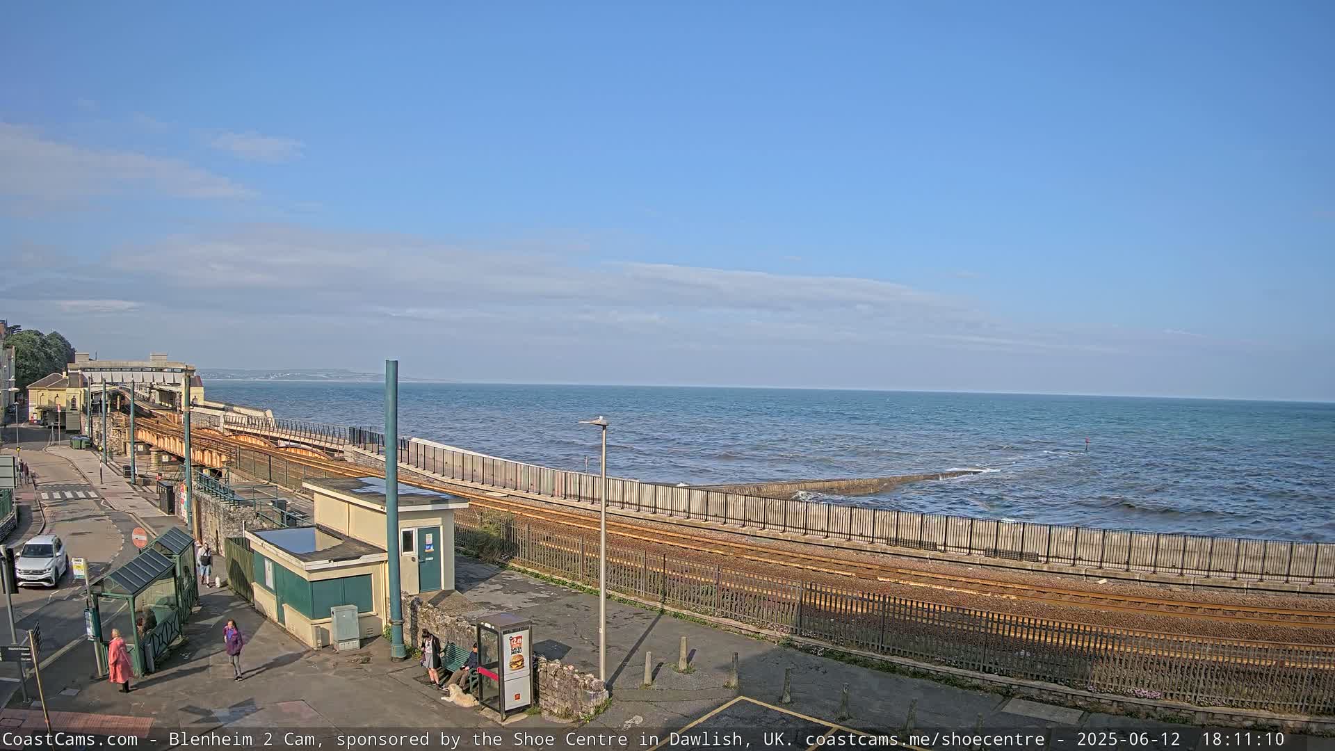 A coastal train track runs alongside a sea wall, with a calm sea visible under a mostly clear sky.