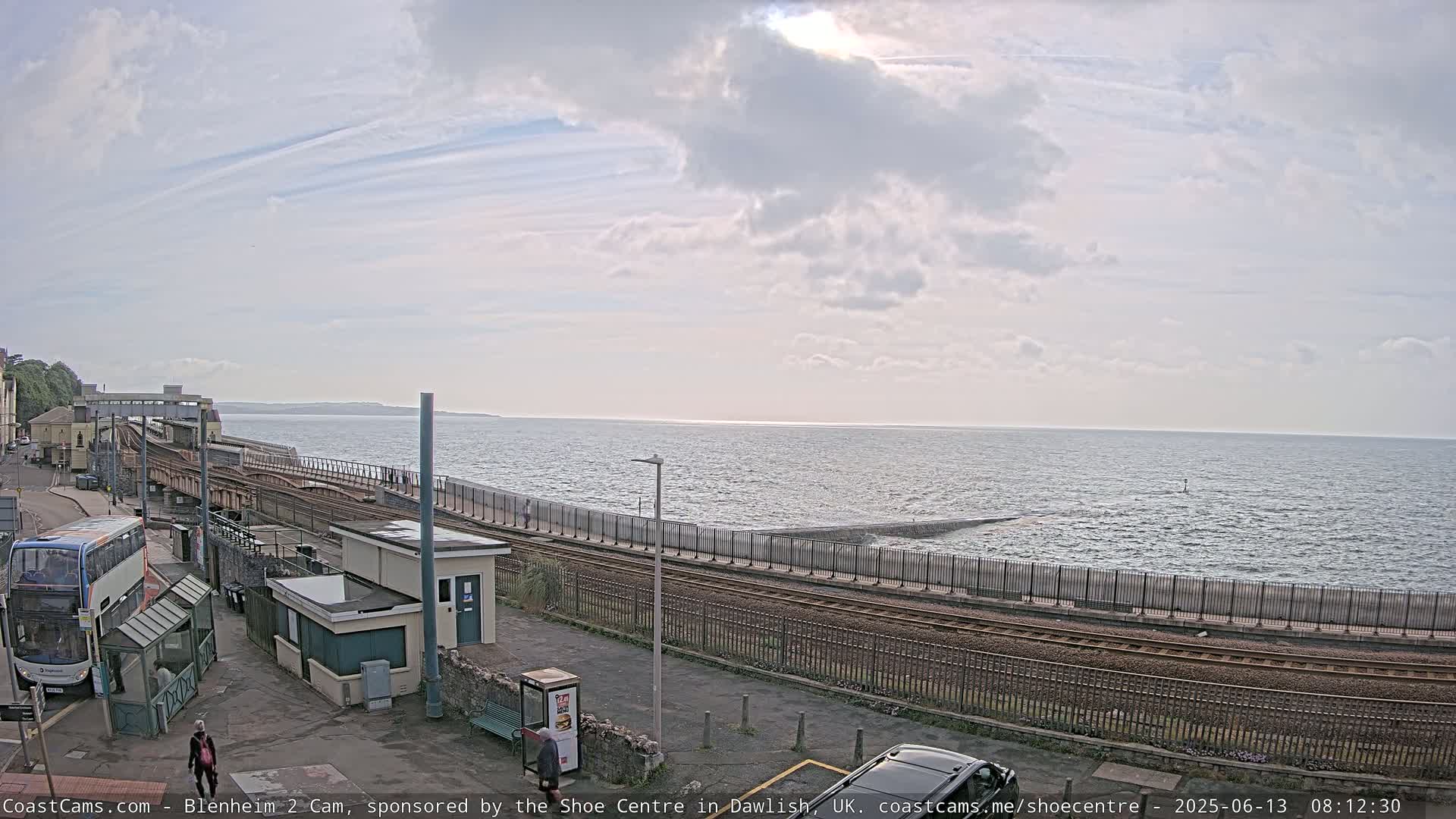 A partly cloudy day shows a train track running alongside a sea wall next to a calm ocean, with a bus and some people visible near a station.