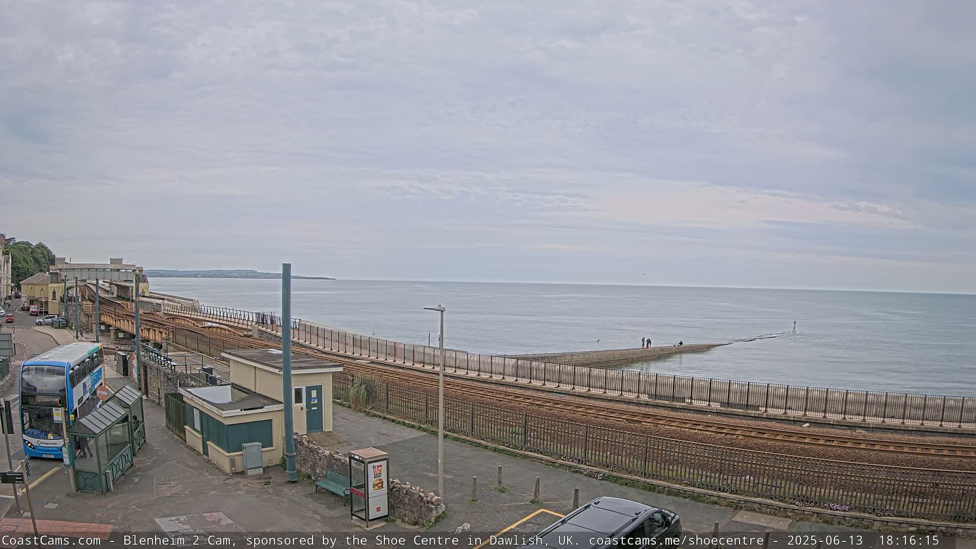 A coastal railway line runs alongside a calm sea under an overcast sky, with a bus and a few people visible near a station.