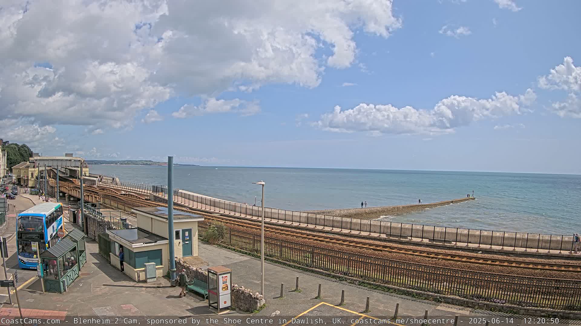 A partly cloudy day reveals a coastal scene with a train track running alongside a seawall, a bus at a station, and calm ocean waters stretching to the horizon.