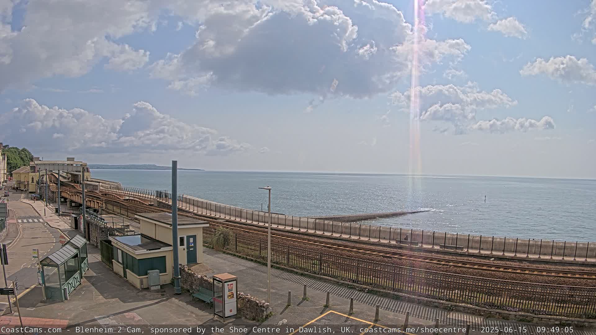 A partly sunny day shows a coastal train station with tracks curving alongside a seawall, overlooking a calm sea.