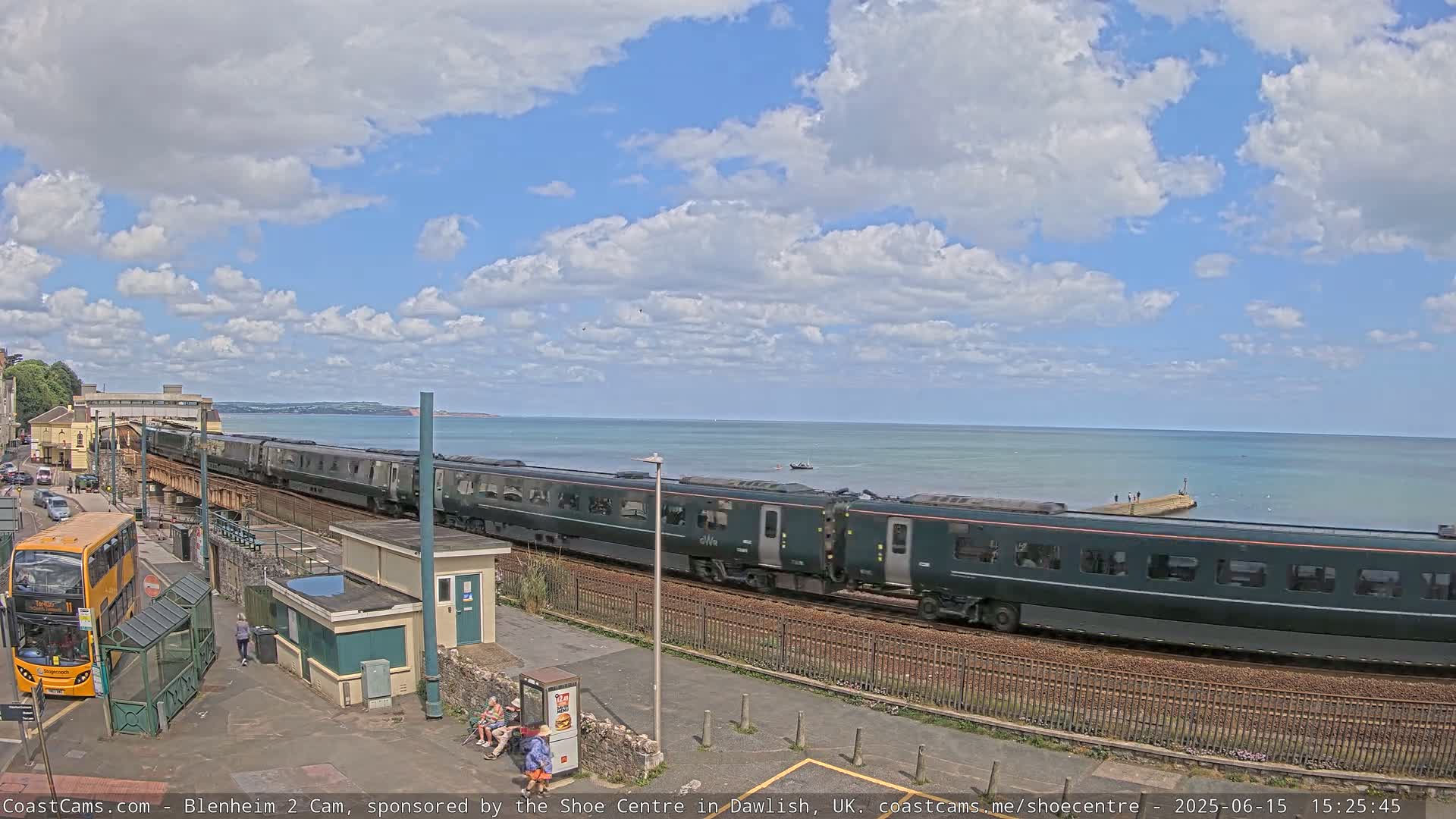 A long dark-green train passes by a seaside train station on a partly sunny day with a mostly blue sky and puffy white clouds.