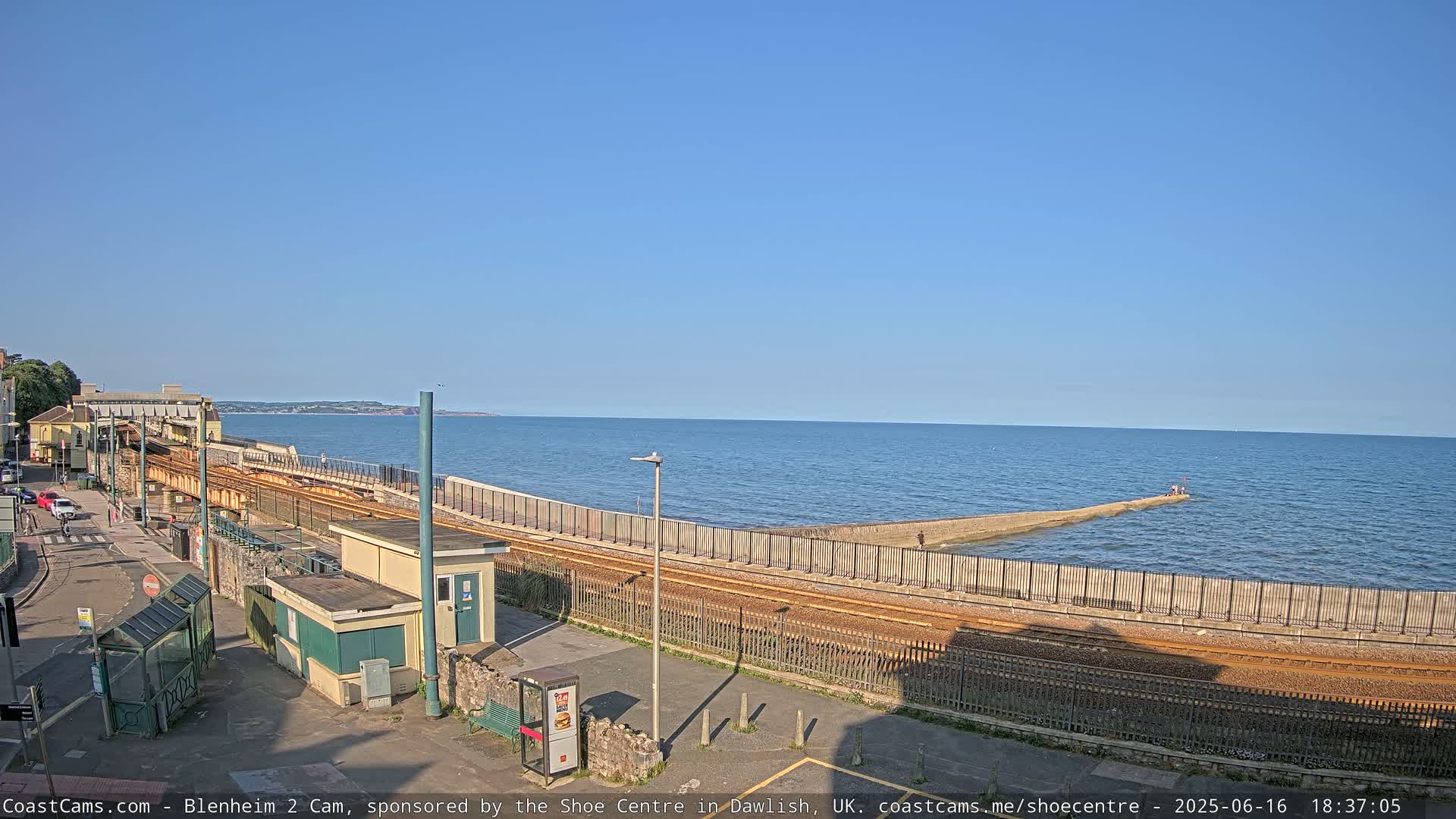 A seaside train track runs alongside a seawall extending into a calm blue ocean under a clear blue sky.