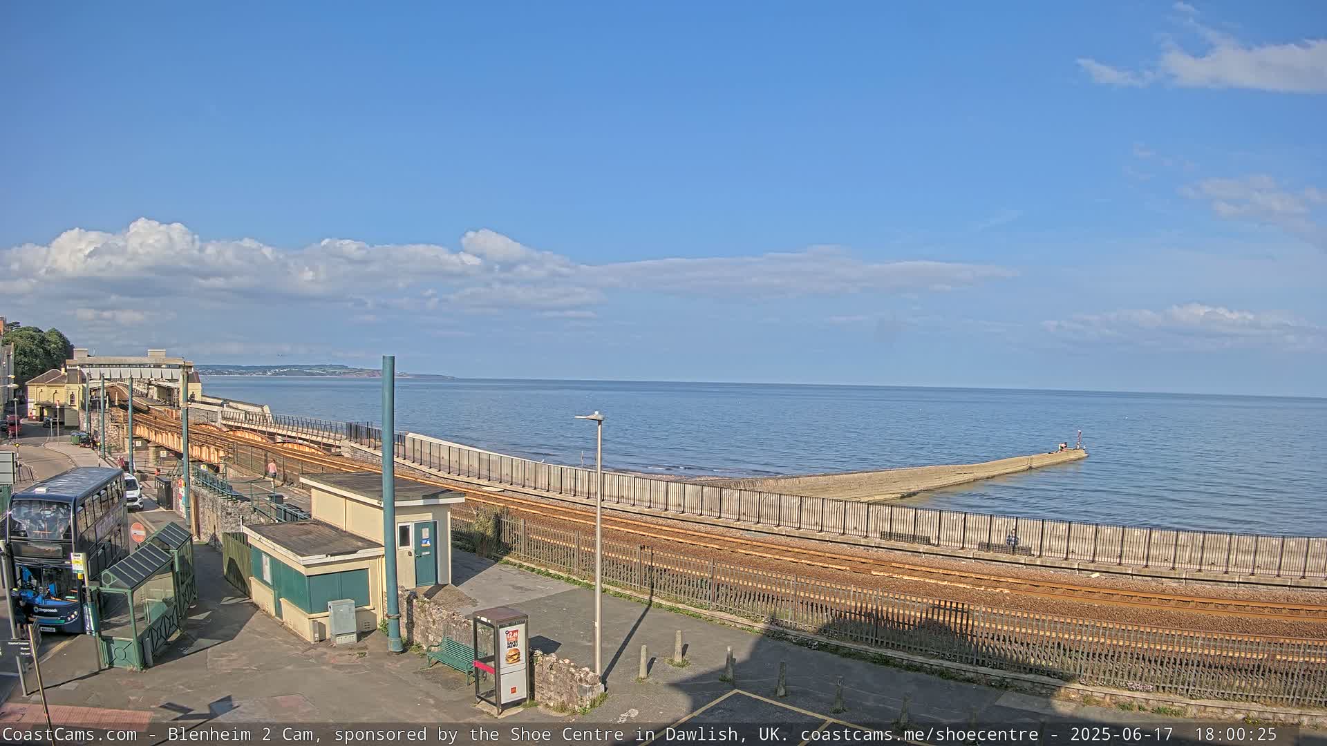 A seaside train track runs along a seawall next to a calm ocean under a partly cloudy sky.
