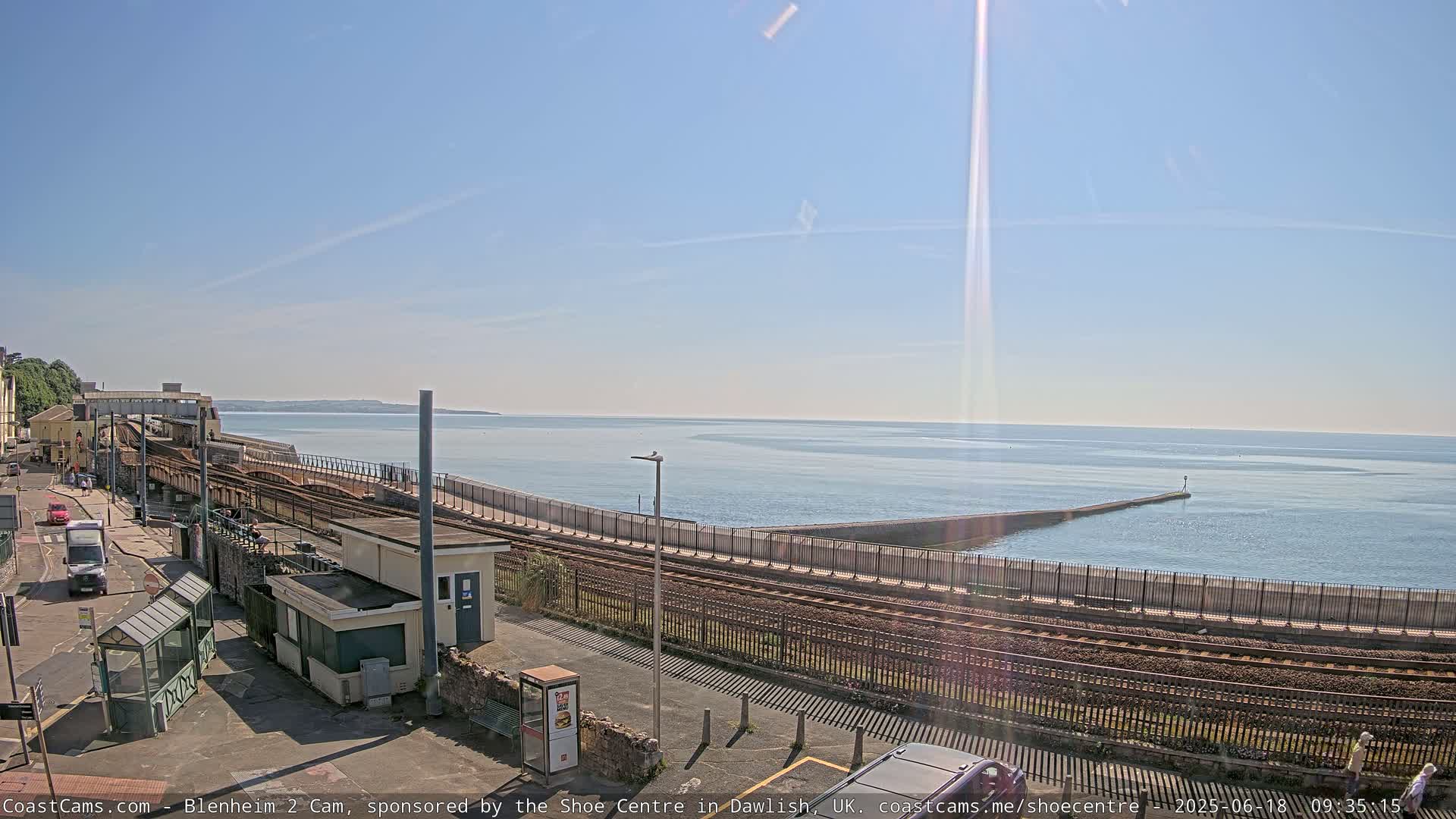 A sunny day overlooks a train track and railway station next to a calm ocean, with a breakwater extending into the water.