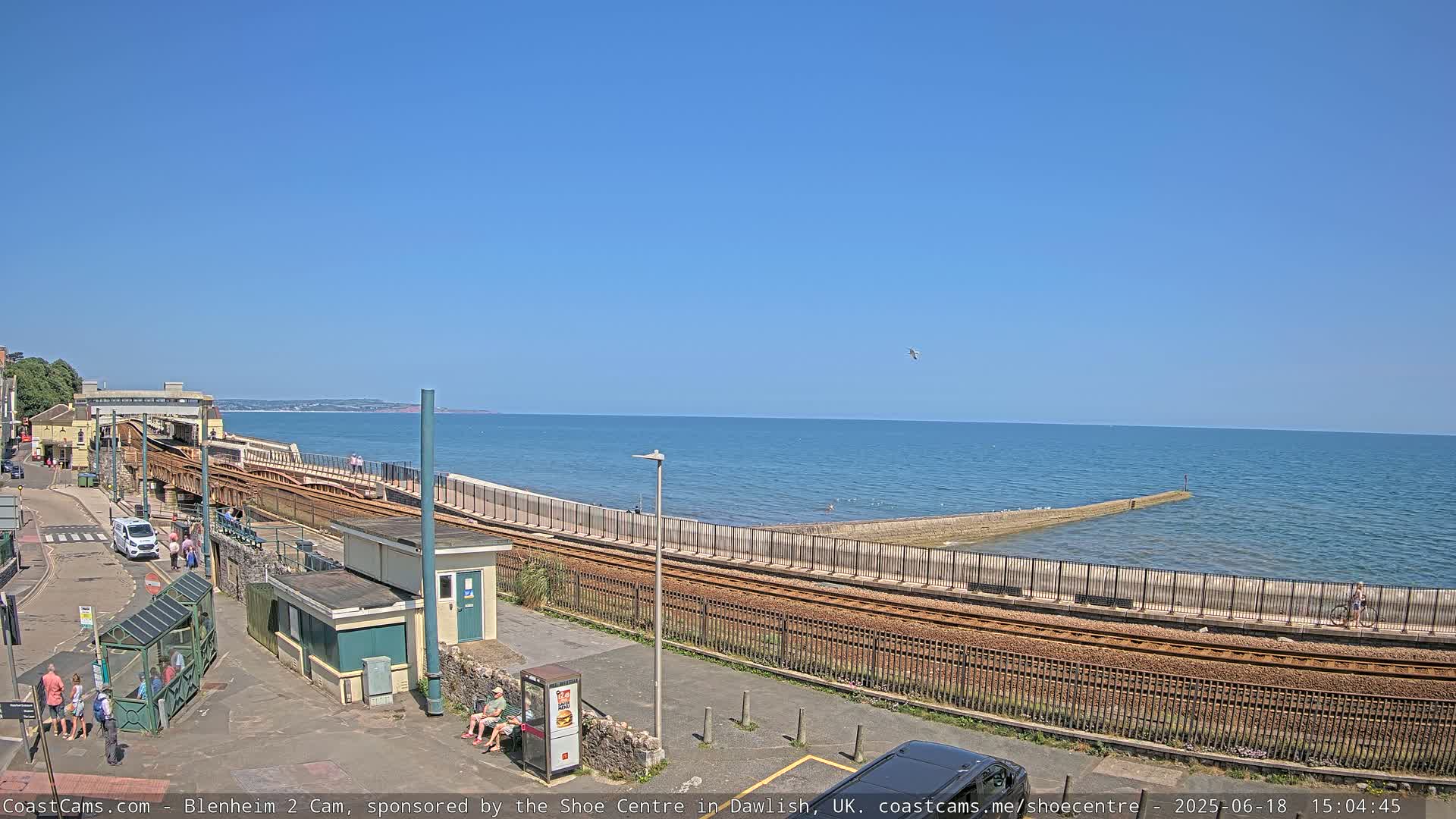 A sunny day at a seaside train station, showing train tracks running alongside a seawall that extends into the calm ocean.