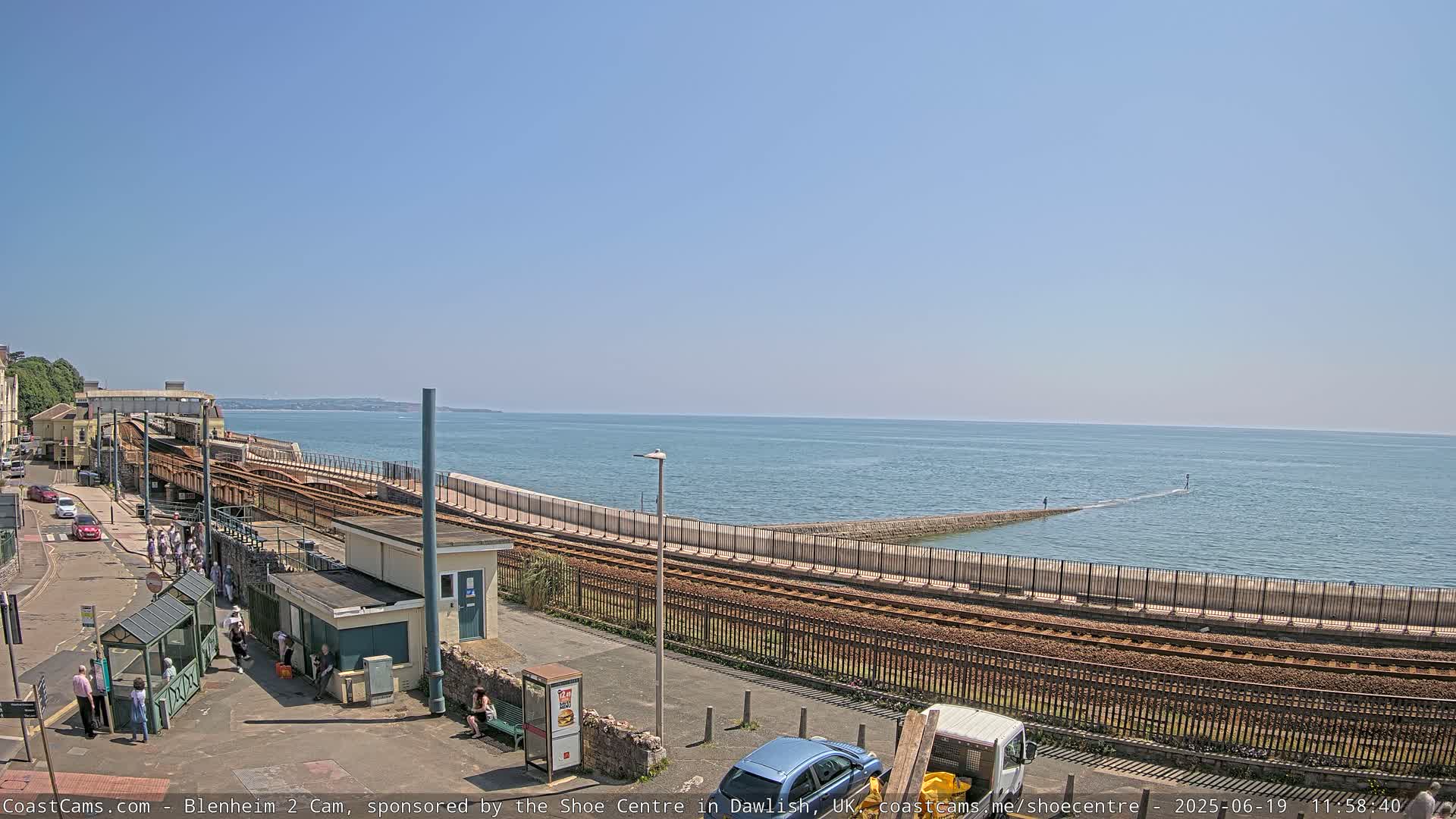 A seaside train track curves along a seawall next to a calm ocean under a clear blue sky.