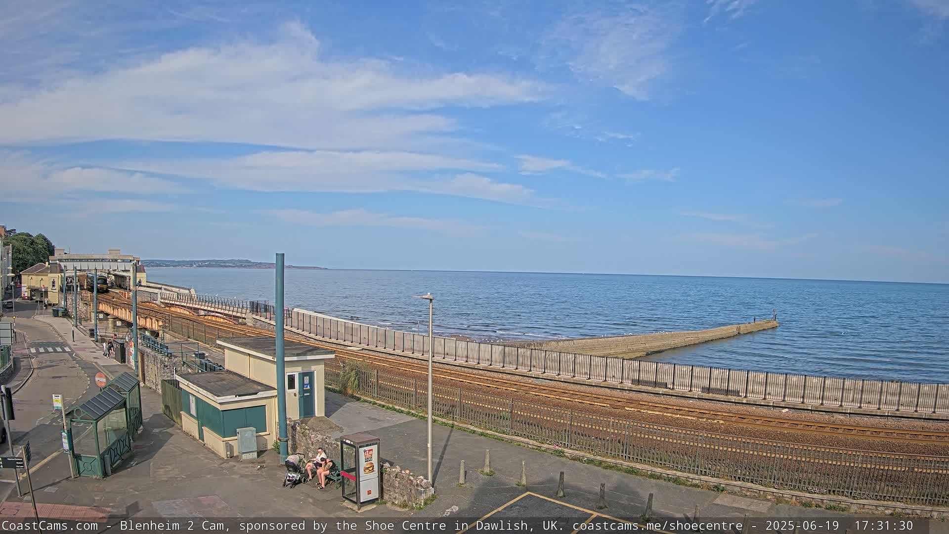 A train track runs alongside a calm ocean under a mostly sunny sky, with a small beach visible in the distance.