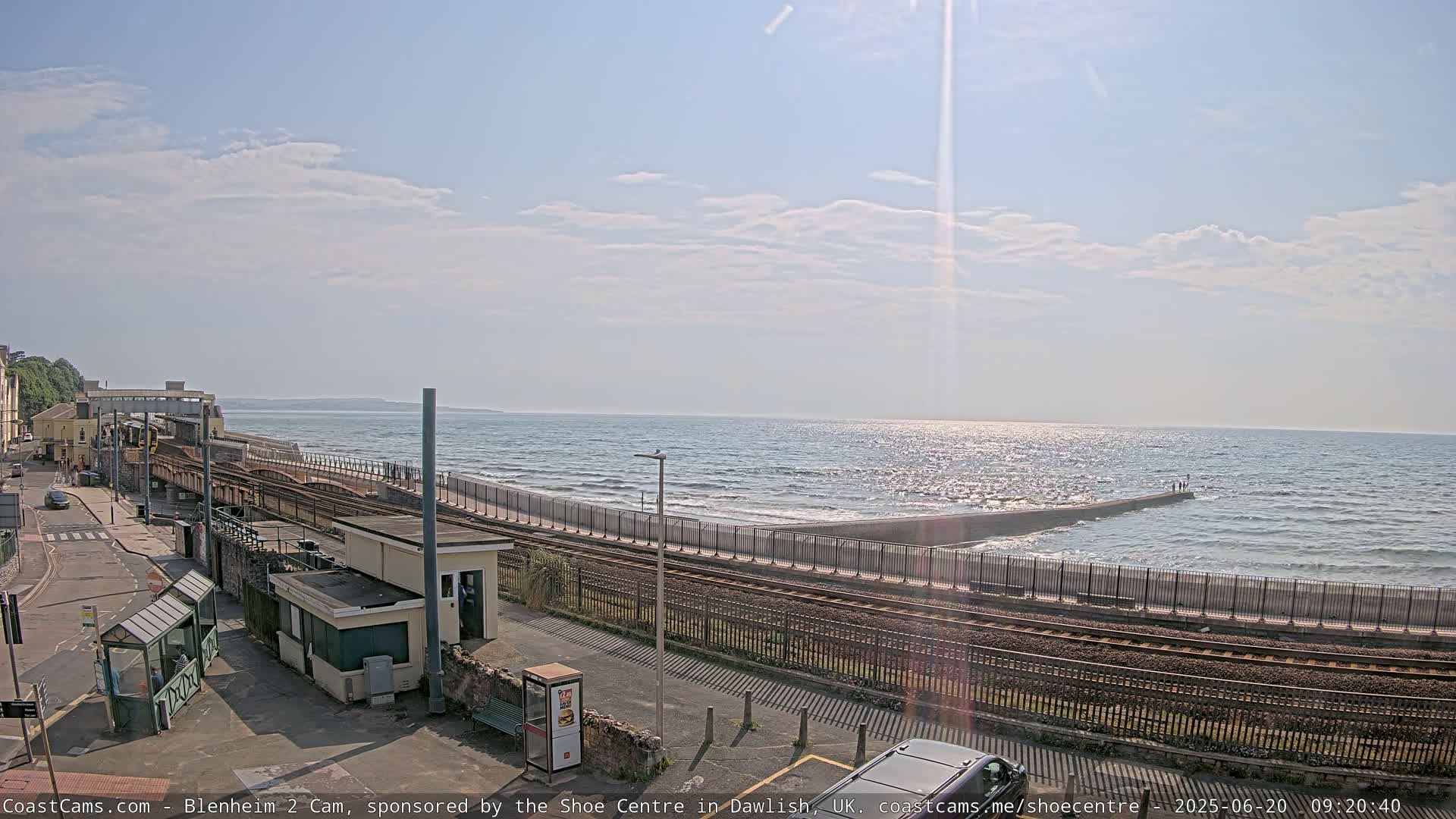 A seaside train station with tracks running alongside a seawall, under a partly sunny sky with some clouds, and a somewhat choppy sea visible in the distance.