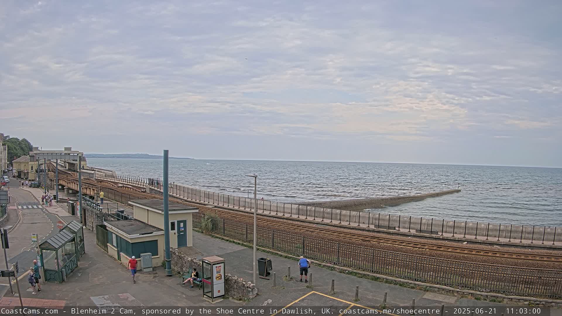 A train track runs alongside a sea wall bordering a calm ocean under a cloudy sky, with several people walking nearby.