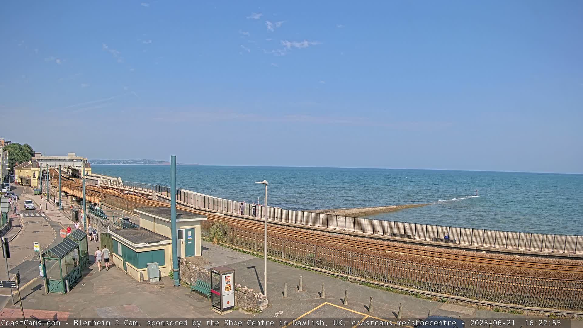 A seaside train track curves along a seawall, overlooking a calm ocean under a clear blue sky.