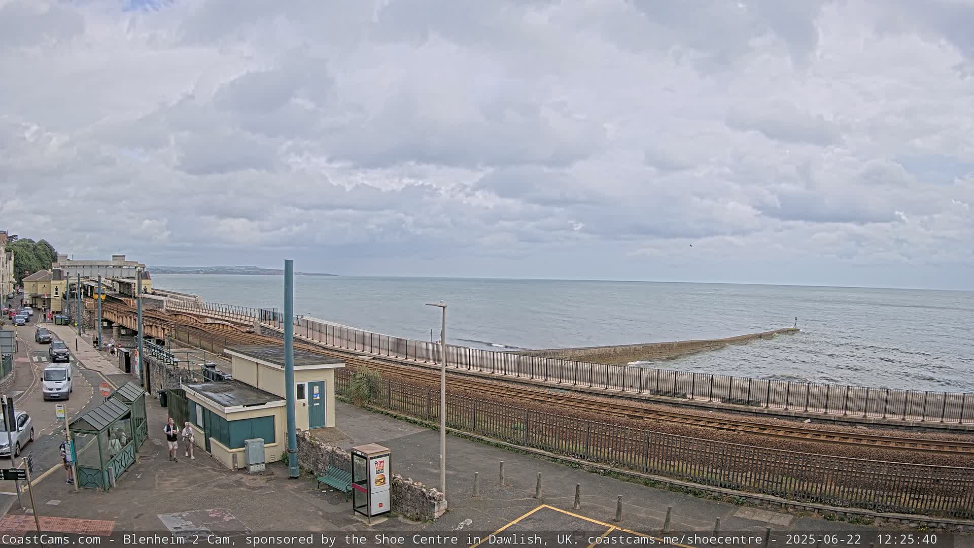 A train track runs alongside a coastline under a cloudy sky, with a small town visible in the background.