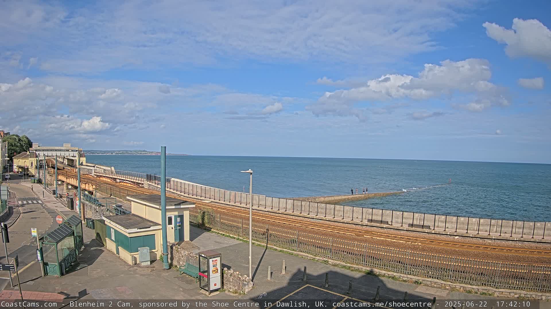 A coastal train track runs alongside a seawall, with a calm sea and partly cloudy sky visible in the background.