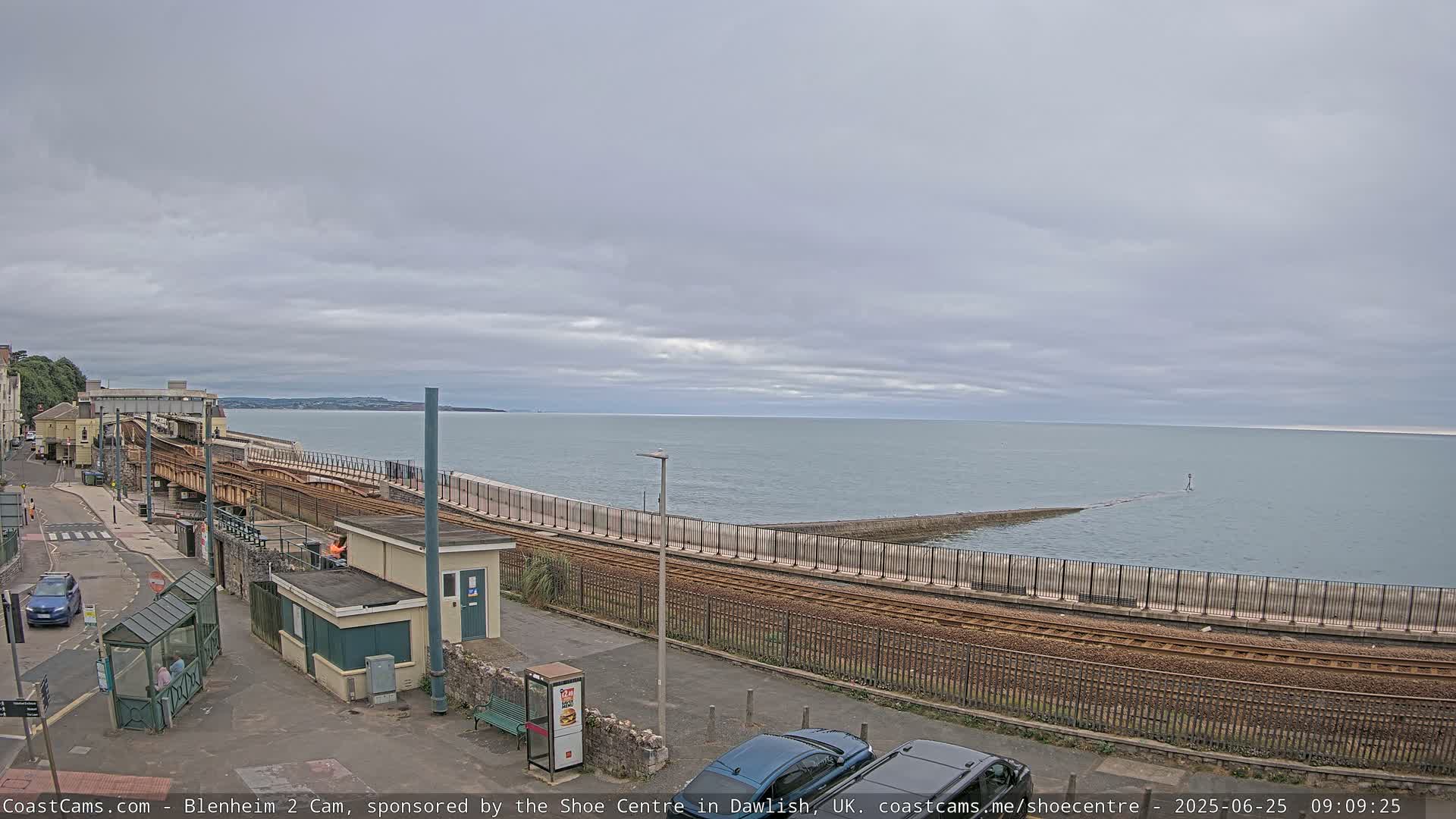 A coastal train track runs alongside a sea wall overlooking a calm sea under a cloudy sky.
