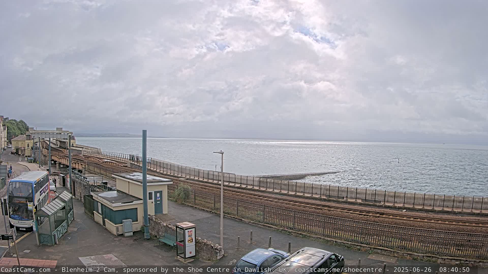 A train track runs alongside a sea wall overlooking a calm, overcast sea, with a bus and several cars visible nearby.