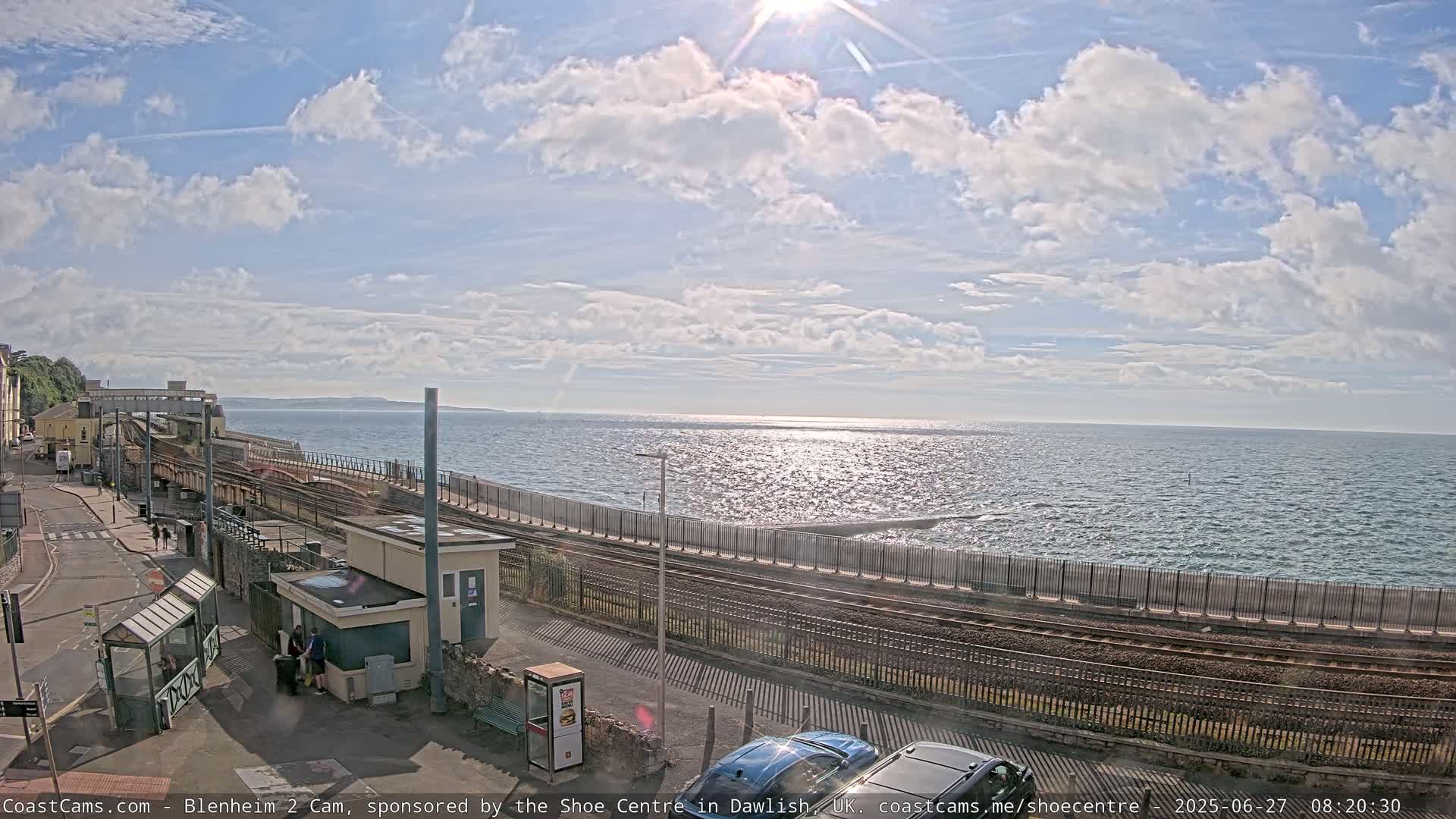 A sunny day with partly cloudy skies overlooks a train track that runs alongside a coastal area with calm ocean waters.