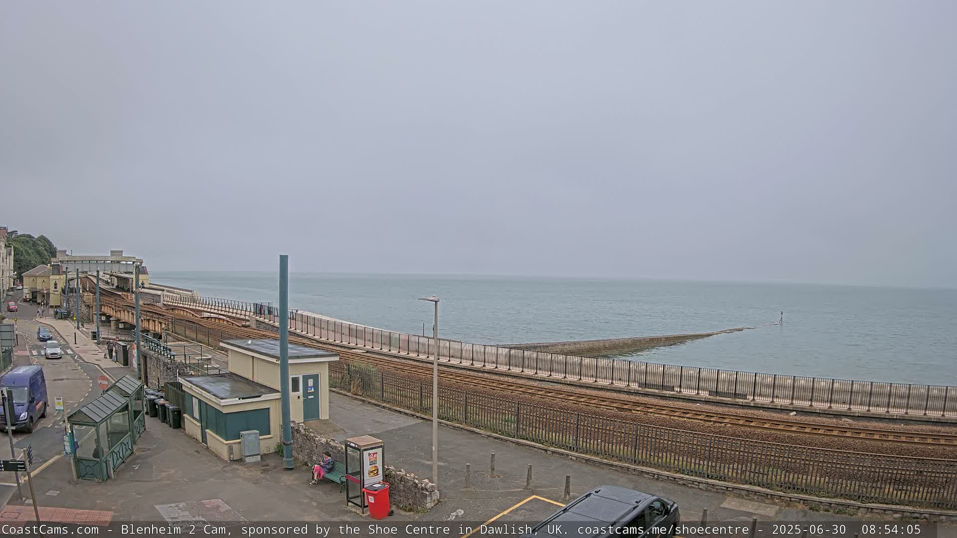 A train track runs alongside a sea wall next to a calm, gray sea under an overcast sky.