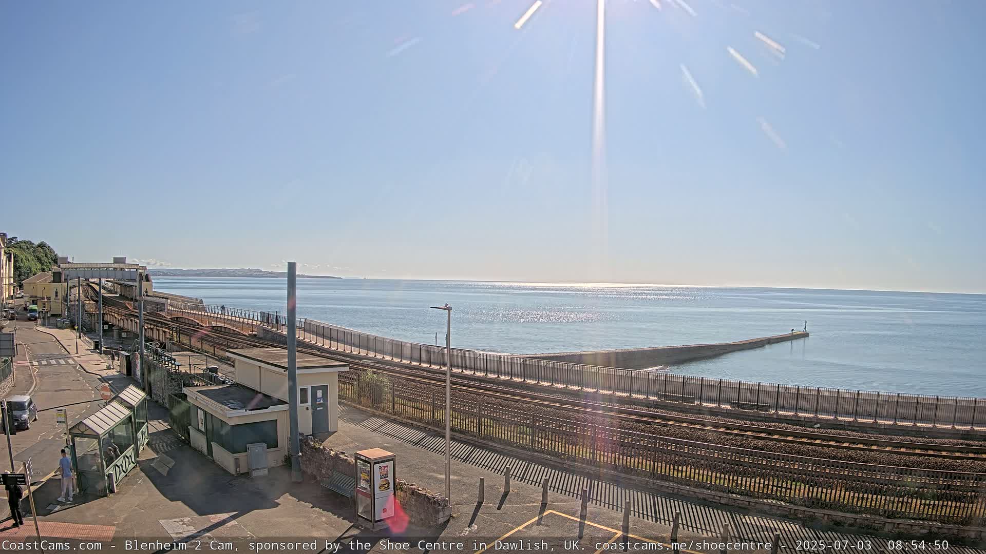 A sunny day reveals a coastal train station, railway tracks, and a seawall extending into a calm ocean.