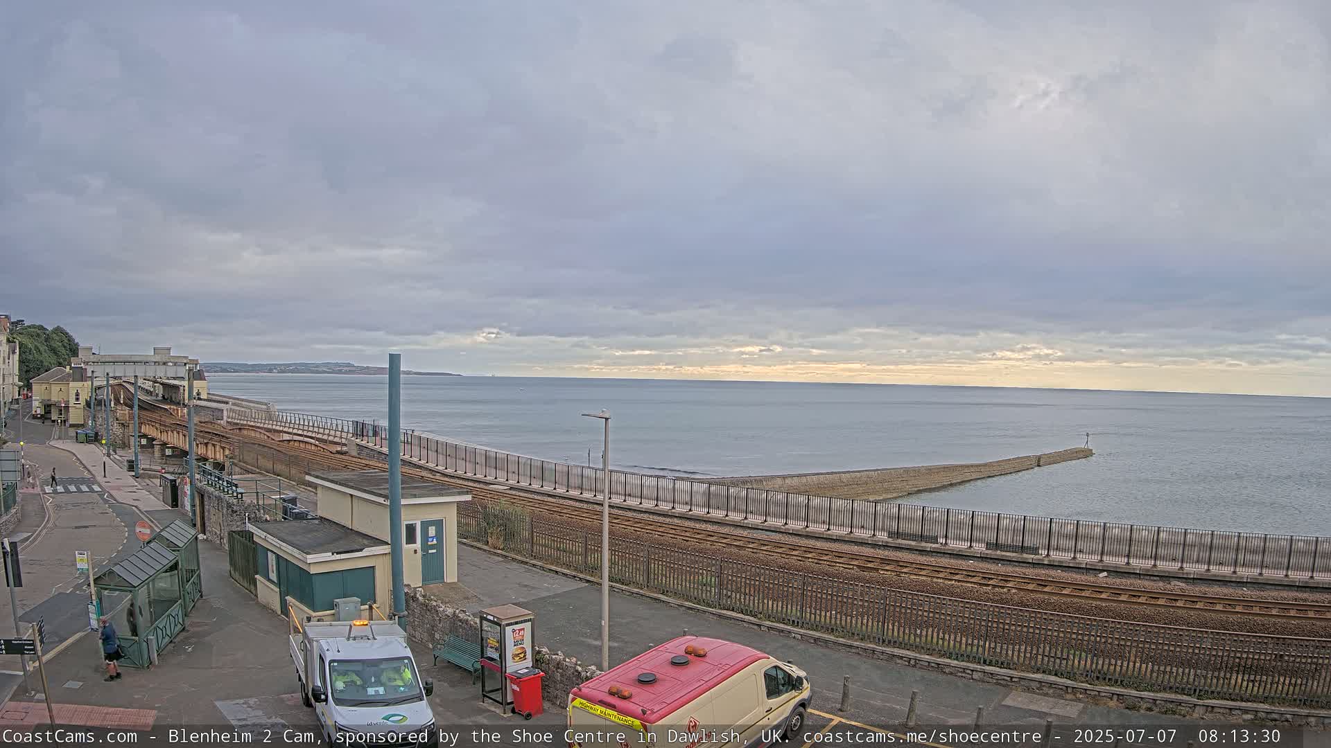A seaside train station with overcast skies and calm waters is seen, with two vans parked nearby and a breakwater extending into the sea.