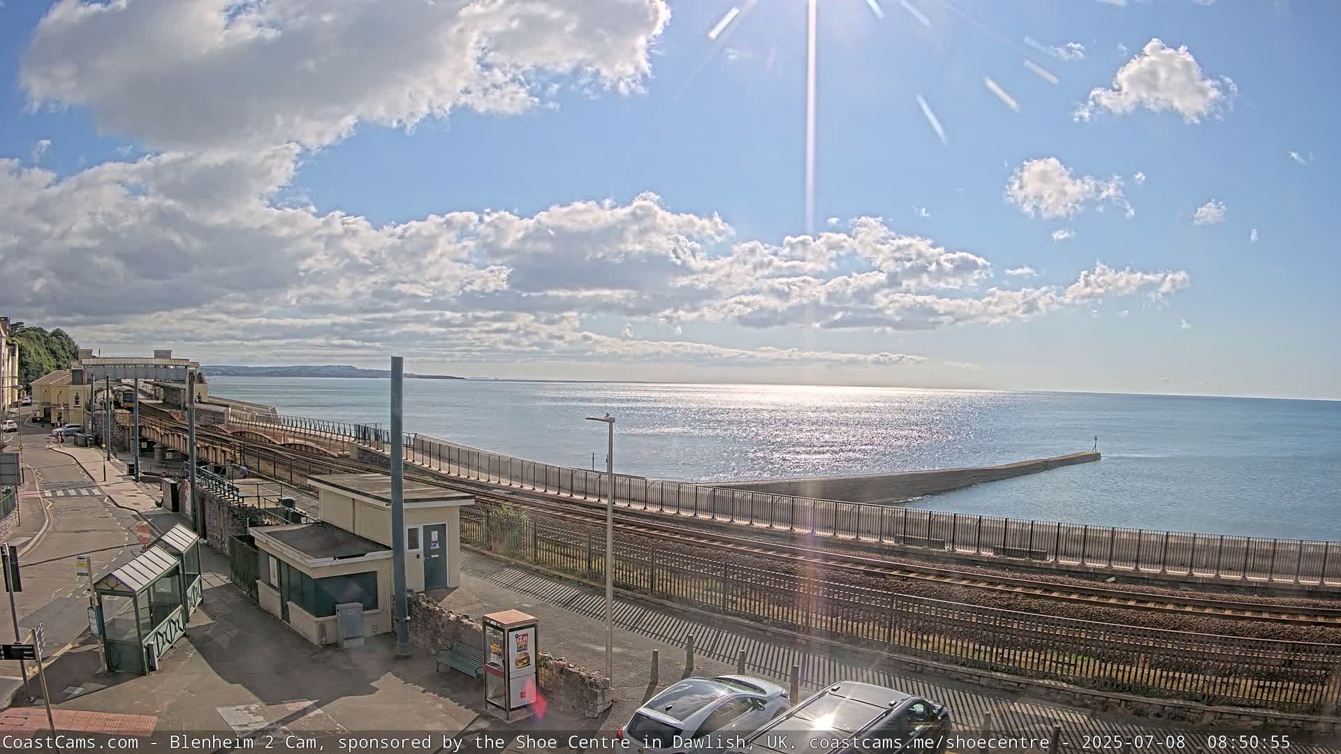 A sunny day with partly cloudy skies overlooks a coastal train station and a calm sea with a breakwater.