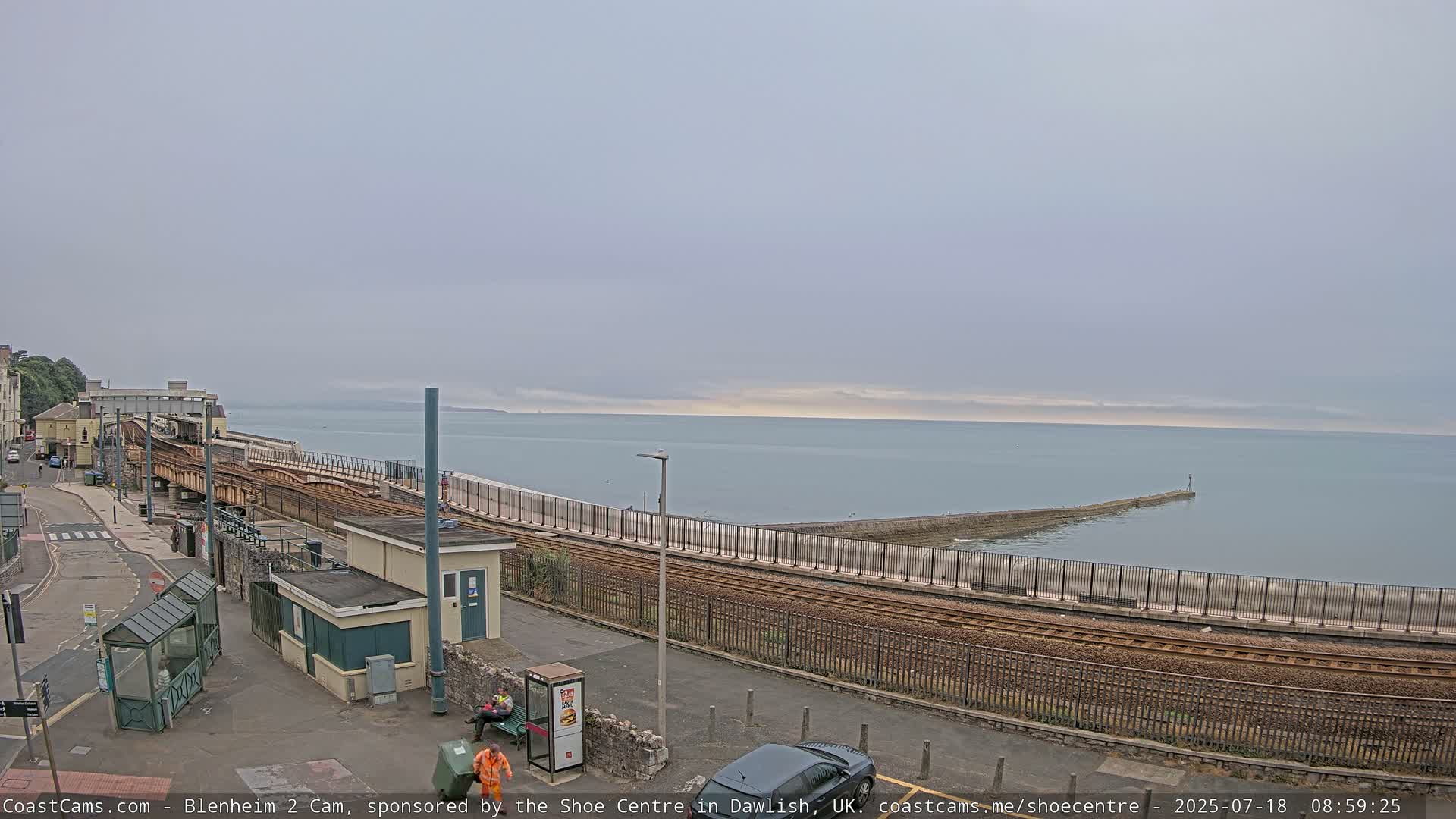 A coastal train track curves alongside a sea wall, overlooking a calm sea under an overcast sky.