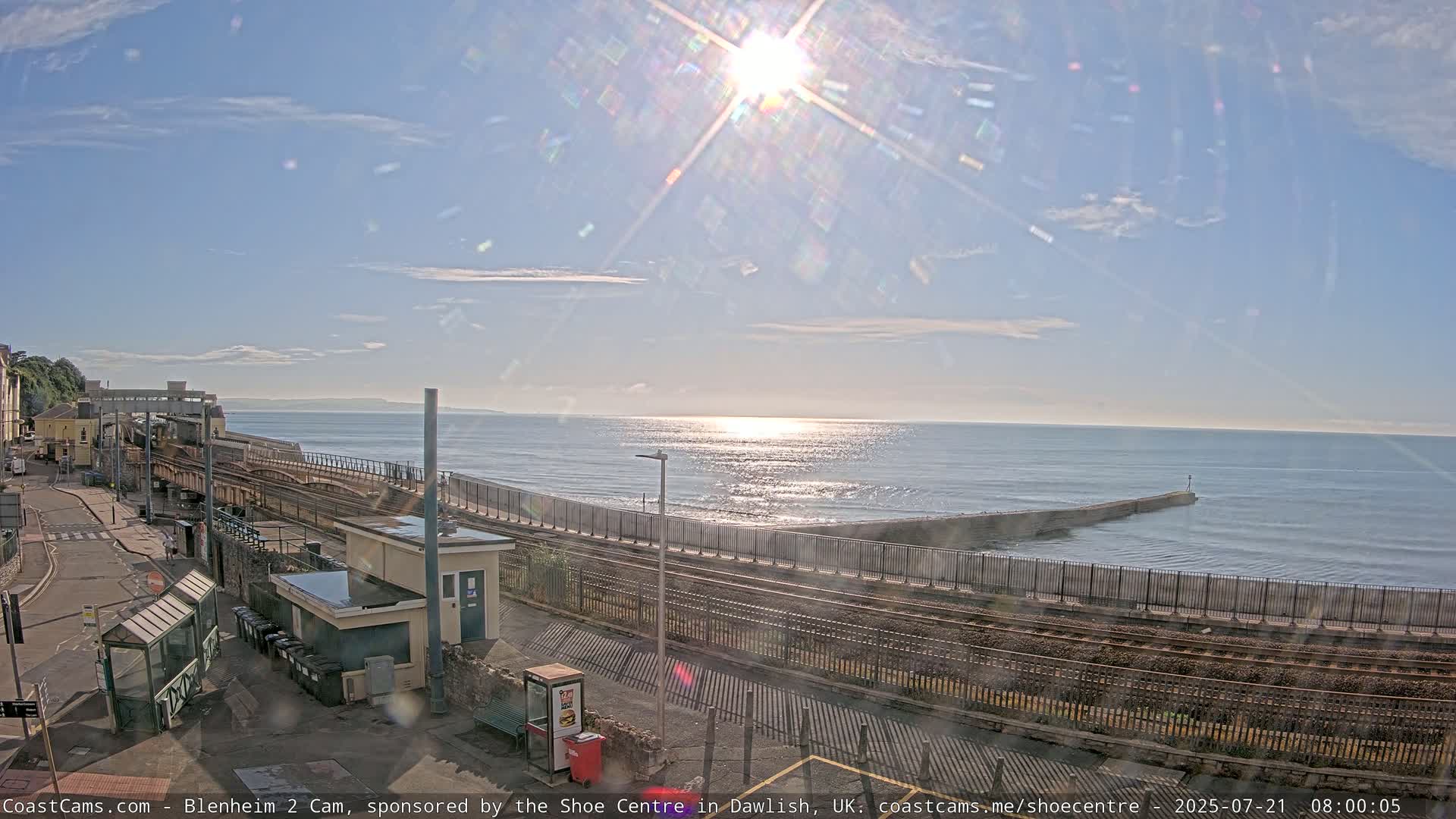 A sunny day shows a train station next to a calm sea with a breakwater, all under a mostly clear sky.