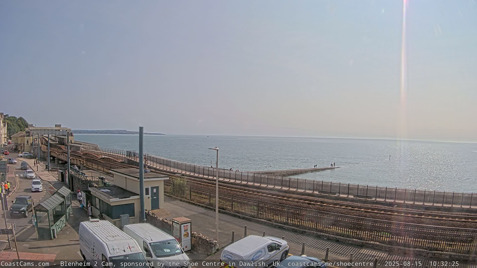A sunny day shows a train station next to a calm sea with a breakwater, all under a mostly clear sky.
