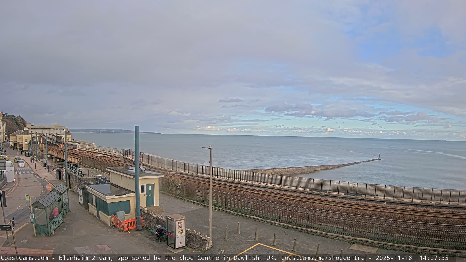 On a bright, clear morning, a coastal scene reveals train tracks and a concrete sea wall bordering a calm ocean with small waves, while a person in an orange jacket scooters along a paved path next to a town built on a hillside.